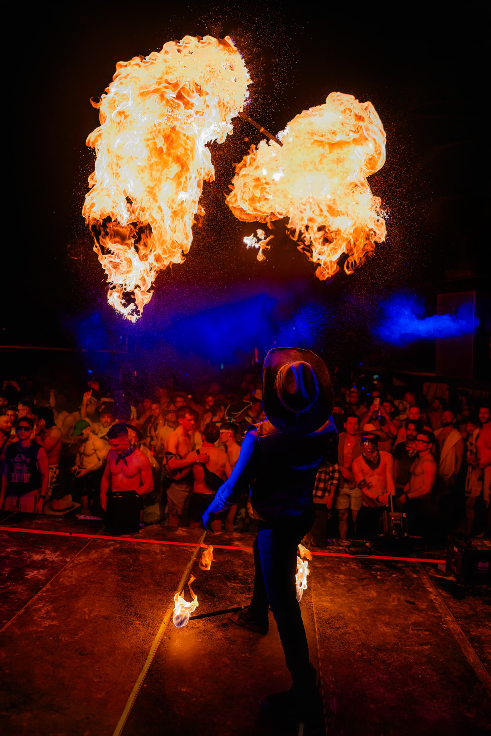 Fire performer in a cowboy hat creates massive fireballs on stage before a large crowd.