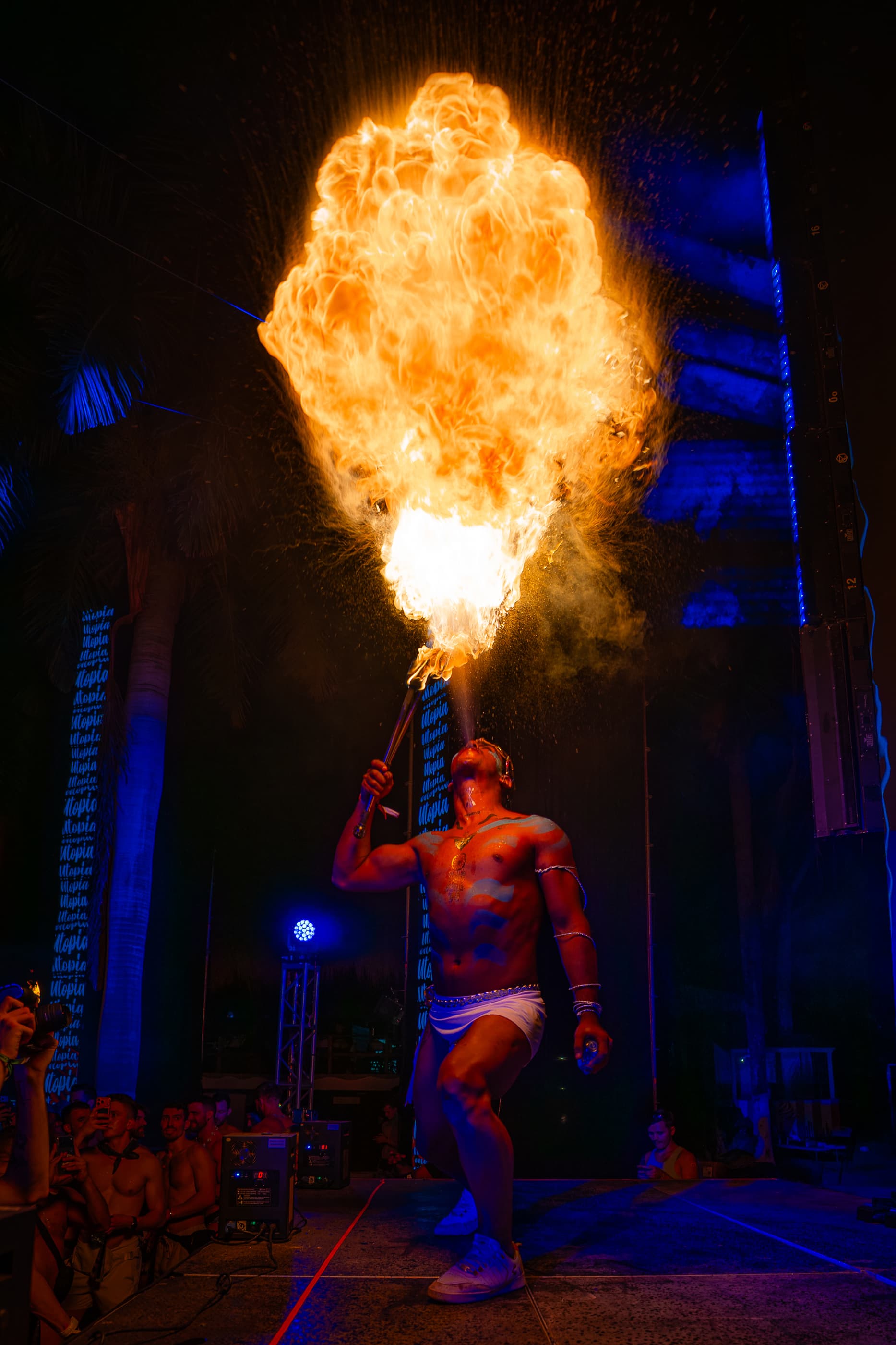 Fire breather with blue body paint blows a massive fireball into the air at night.