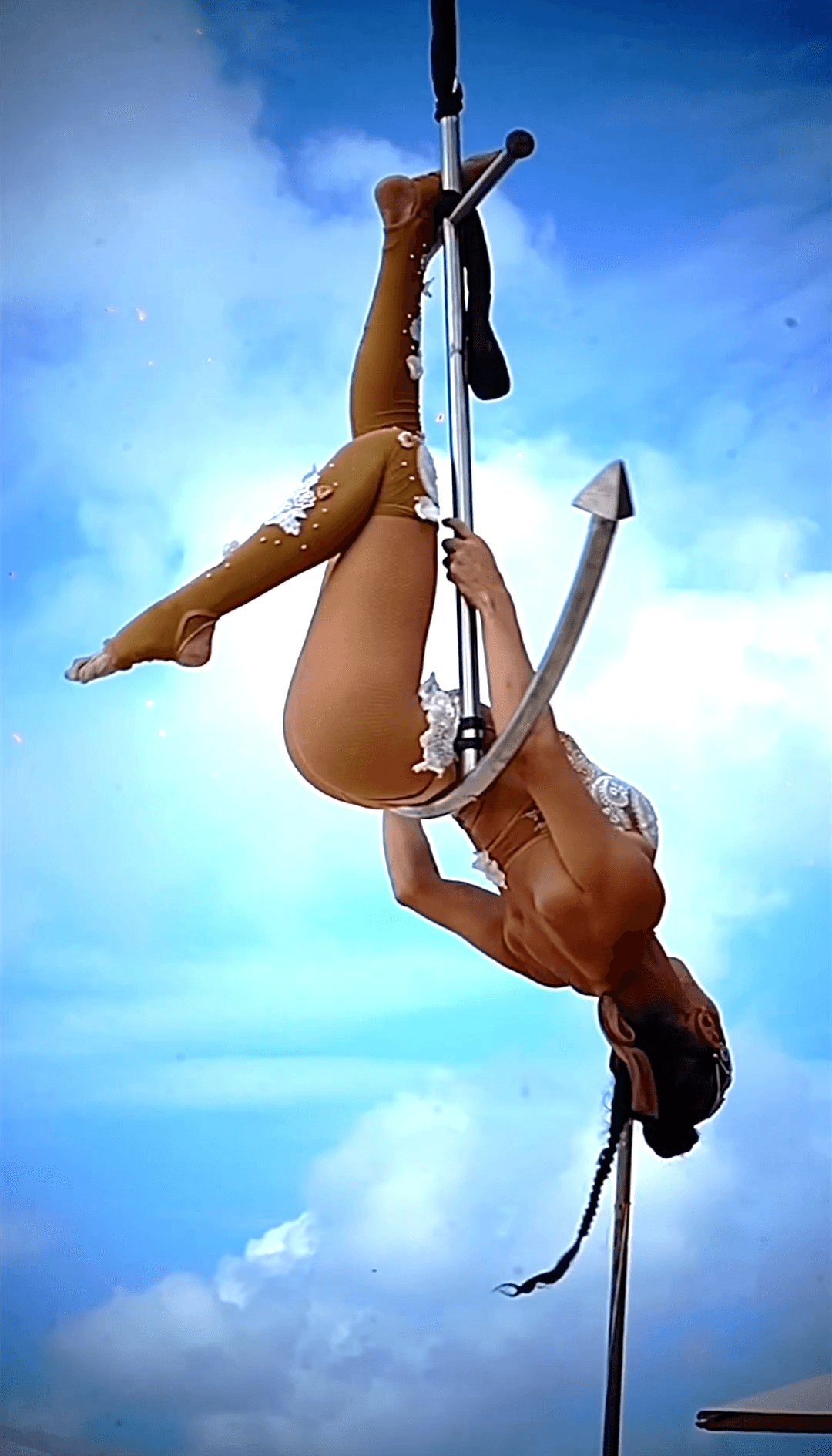 Upside-down aerialist in tan lace costume performs on an anchor-shaped pole against blue sky.