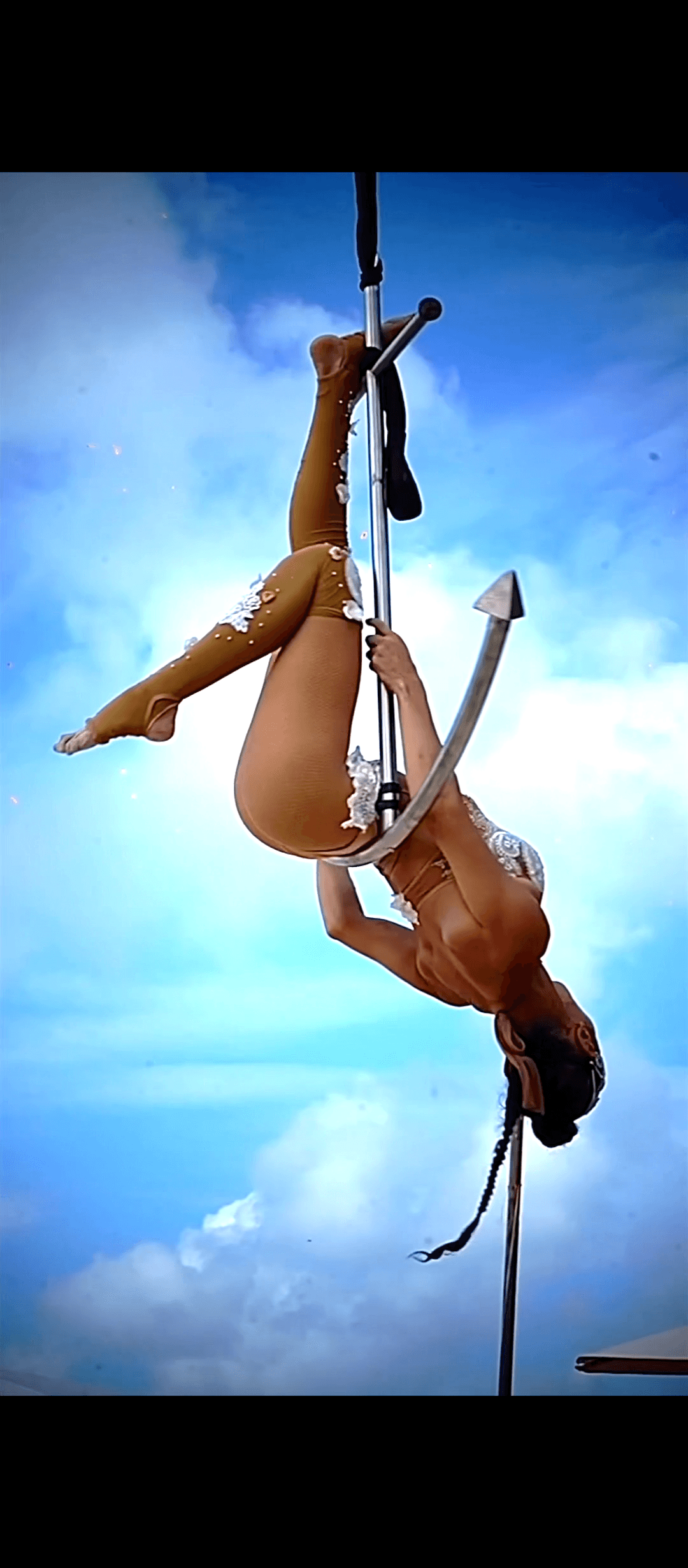 Aerialist hanging upside down from an anchor-shaped pole against a bright blue sky.