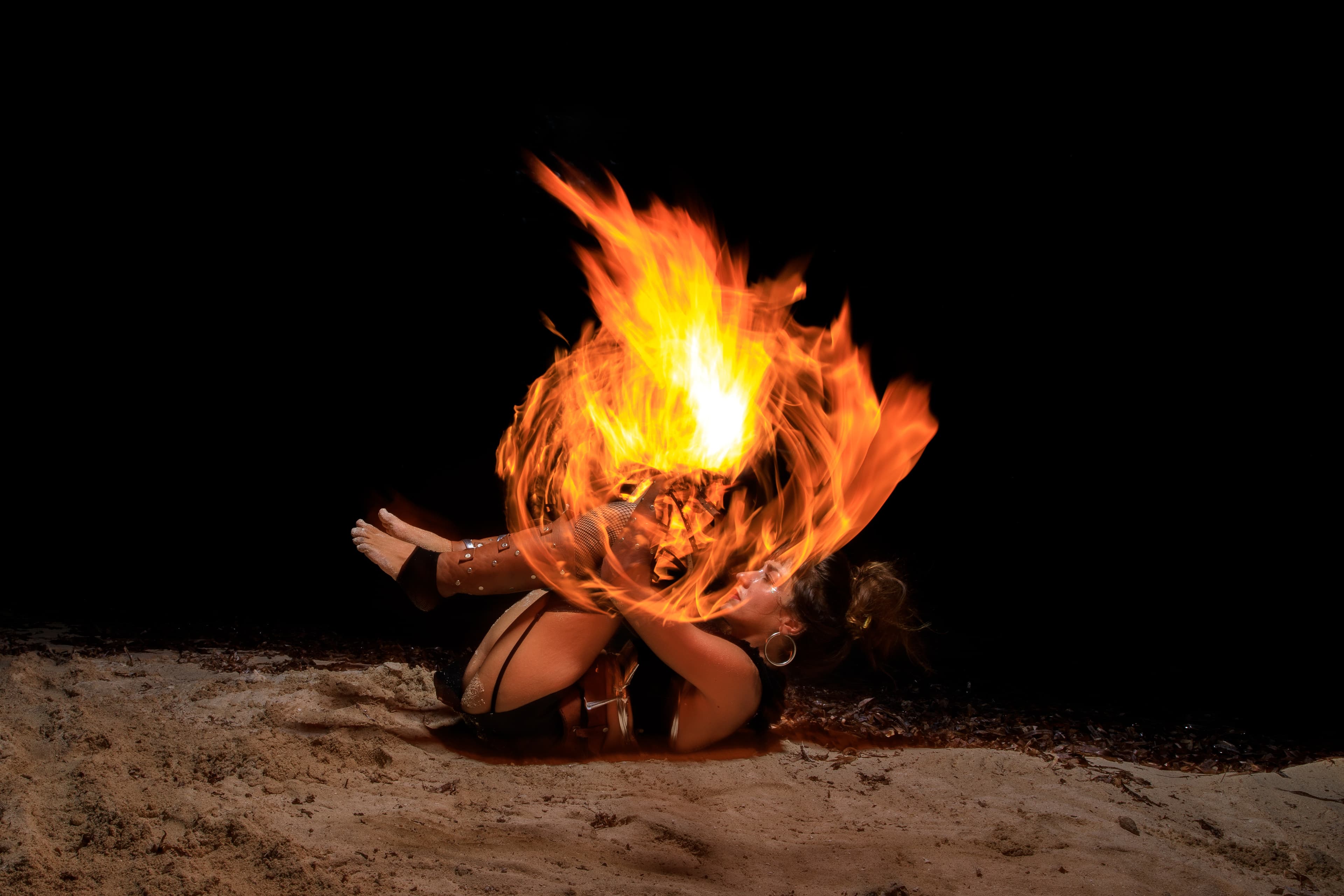 Female fire performer curled on a beach at night with a large, swirling orange flame.